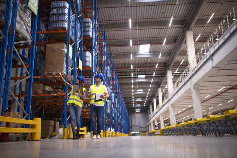 Shot of two workers walking through large warehouse center, observing racks with goods and planing distribution to the market